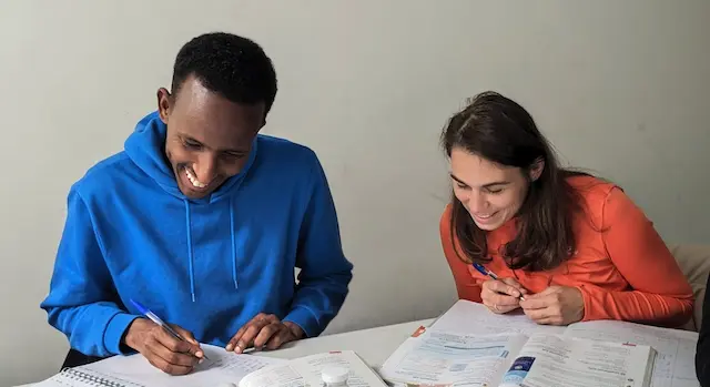 Hassan et Yulia travaillant côte à côte et en souriant sur des exercices dans un manuel scolaire.